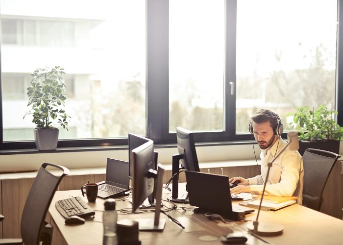 A businessman sits at a desk using multiple computers and a headset in a well-lit modern office.